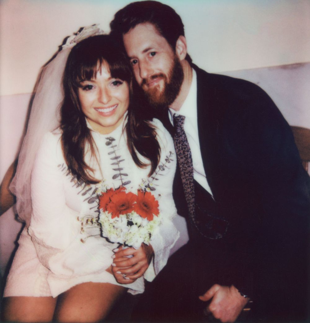 A newly married couple poses with red and white flowers in casual wedding attire.