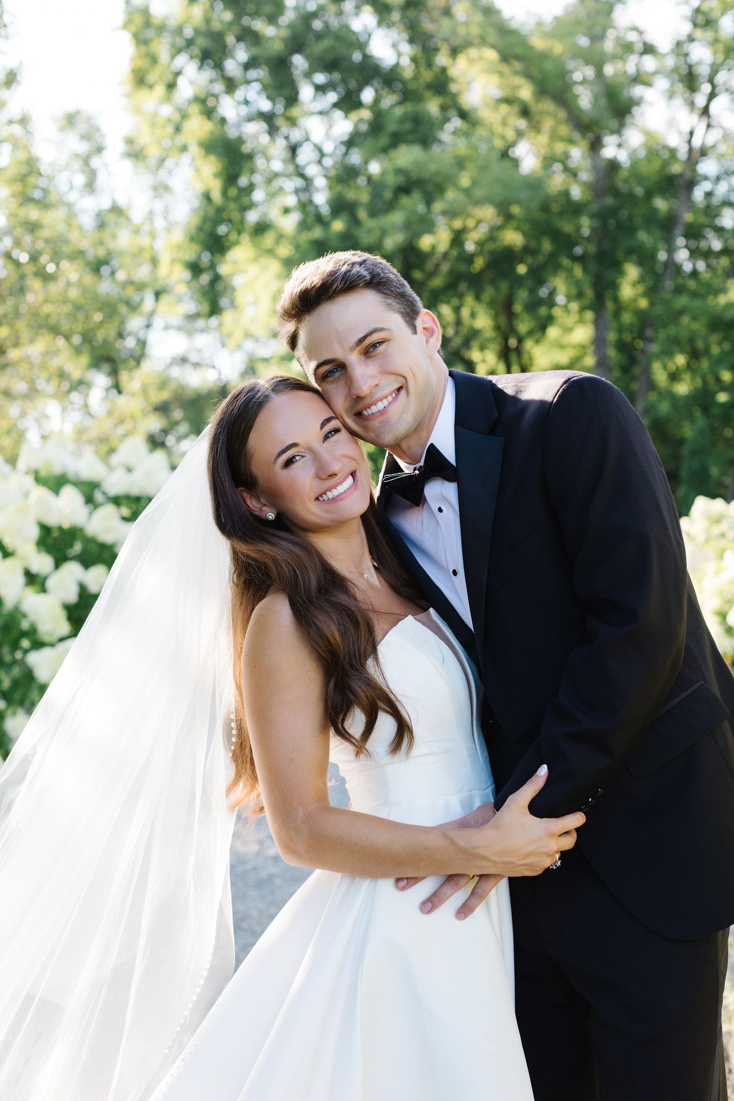 A smiling couple in a white wedding dress and black tuxedo embrace outdoors on their special day.