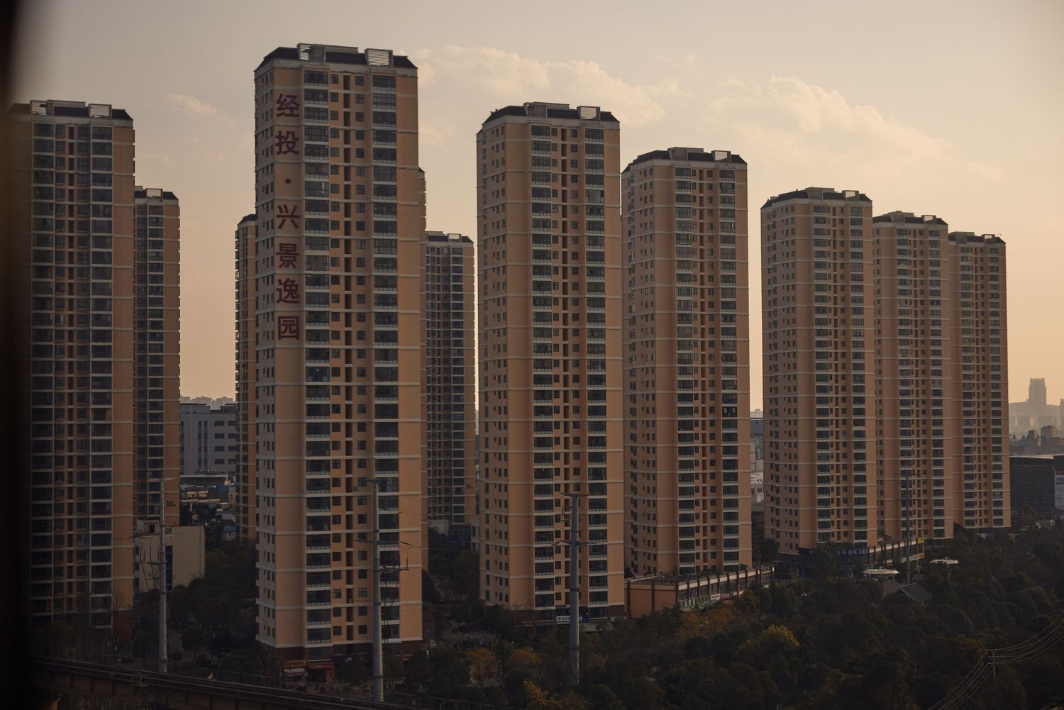 A row of tall residential apartment towers with beige facades stand against a dusky sky in an urban setting.