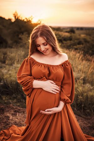 A woman in a flowing rust-colored dress cradles her baby bump during golden hour sunset in a serene outdoor setting.