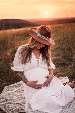 Pregnant woman in white dress and brown hat sits on blanket during golden sunset in field of tall grass.