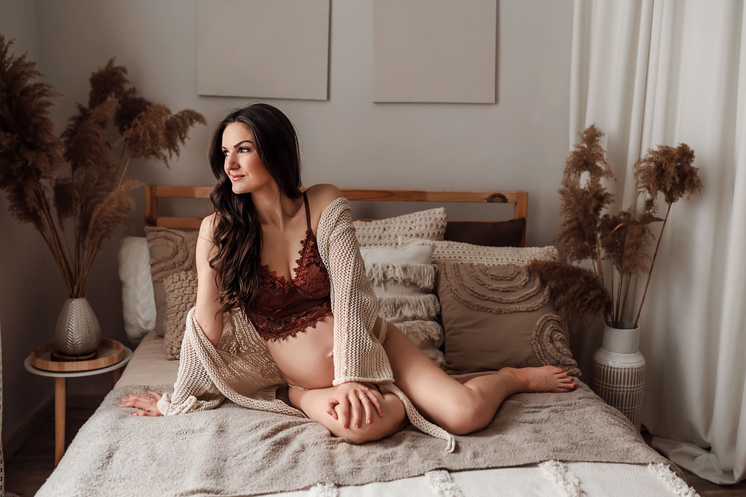 Relaxing on a cozy bed with dried pampas grass decor, wearing a burgundy top and beige cardigan in soft natural light.
