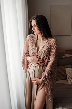 Expecting person in pink knit cardigan and beige dress stands by window in intimate maternity portrait.