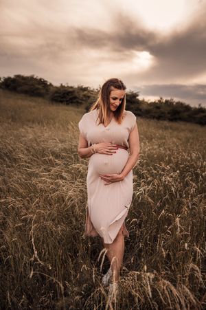 A pregnant figure in a pink dress stands in a golden wheat field at sunset, gently cradling their belly.