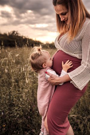 A child kisses an expectant mother's belly in a field at sunset while wearing a pink outfit.