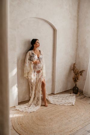 Expectant mother in flowing lace robe poses against white wall with natural light and boho decor elements.