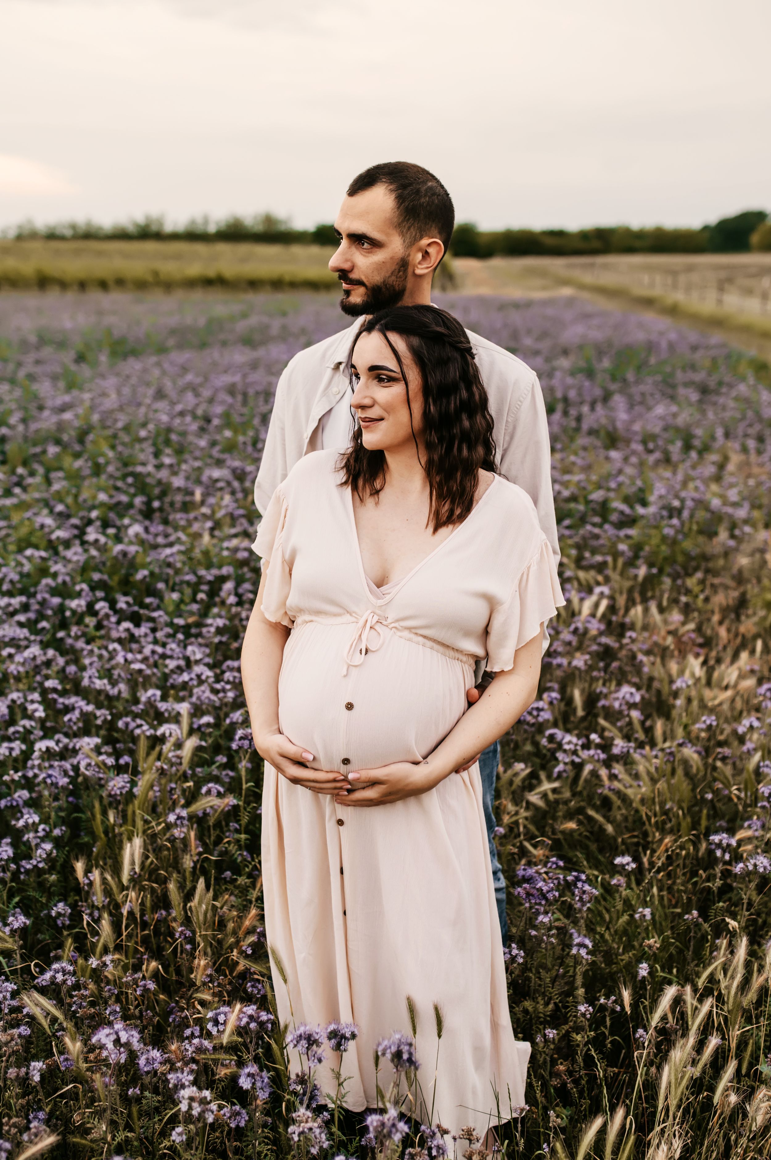 A couple stands together in a blooming purple flower field at sunset wearing light colored clothing.