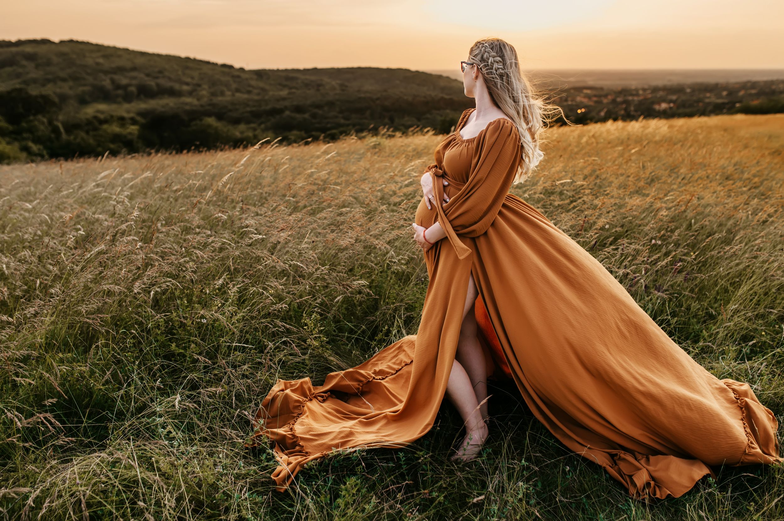 A flowing orange dress billows in the wind against a golden sunset over grassy meadow during an outdoor photoshoot.