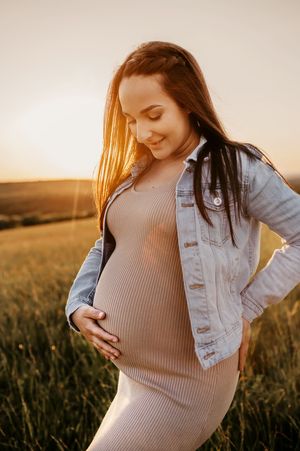 Expectant mother in ribbed dress and denim jacket stands in golden sunlit field at sunset, touching her belly.