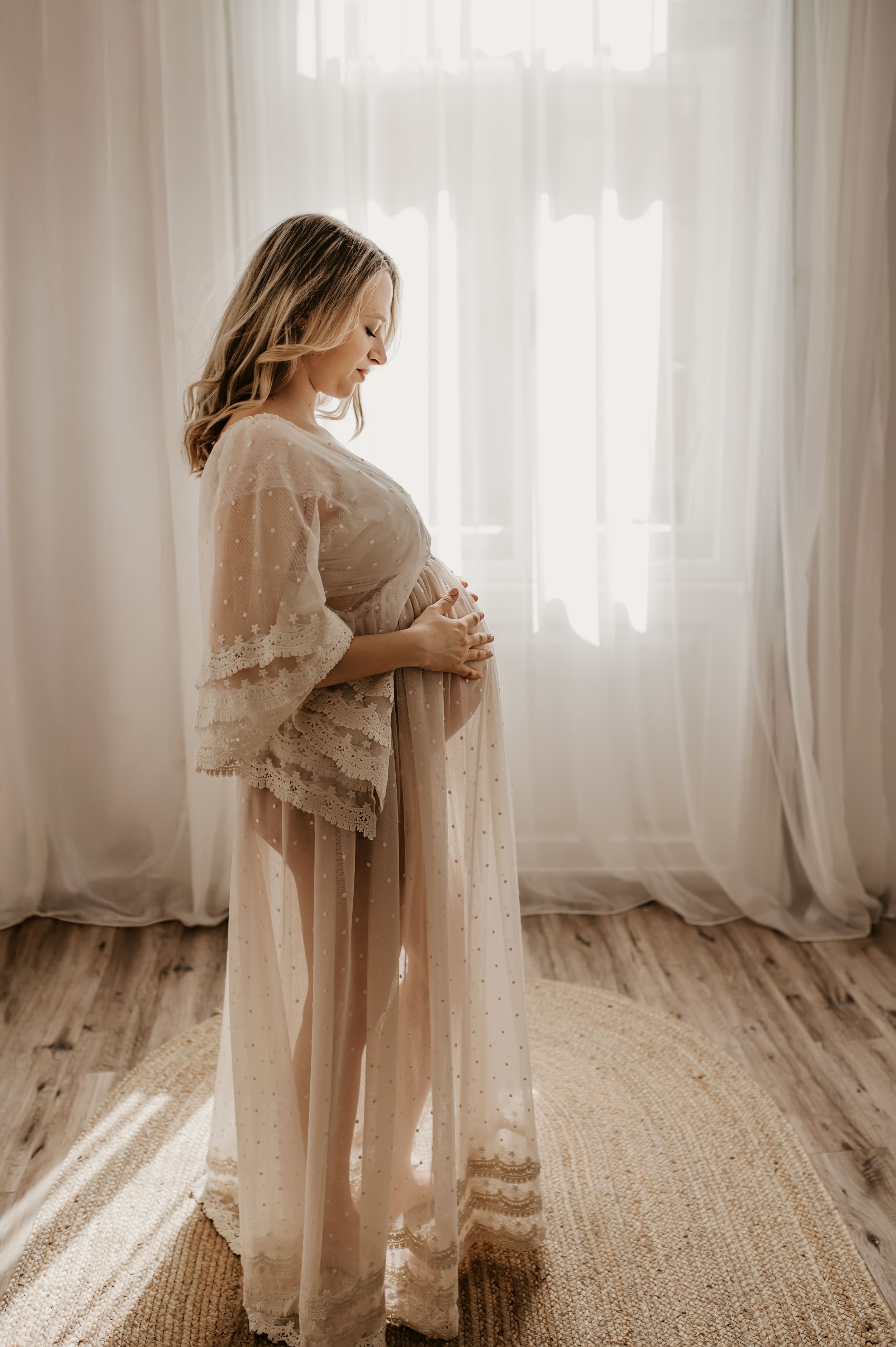 Pregnant woman in sheer lace maternity gown cradling baby bump by sunlit white curtains.