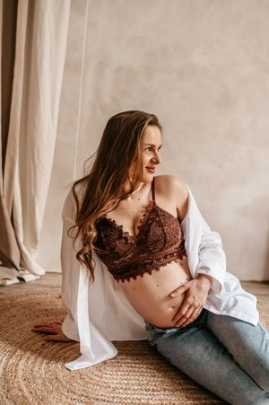 A woman in a burgundy lace bralette and white shirt lounges on a jute rug in casual maternity boudoir pose.