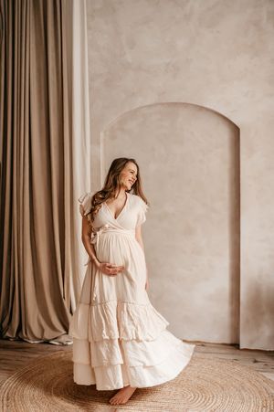 Expectant woman in flowing white tiered maxi dress stands gracefully against beige wall with arched alcove and curtains.