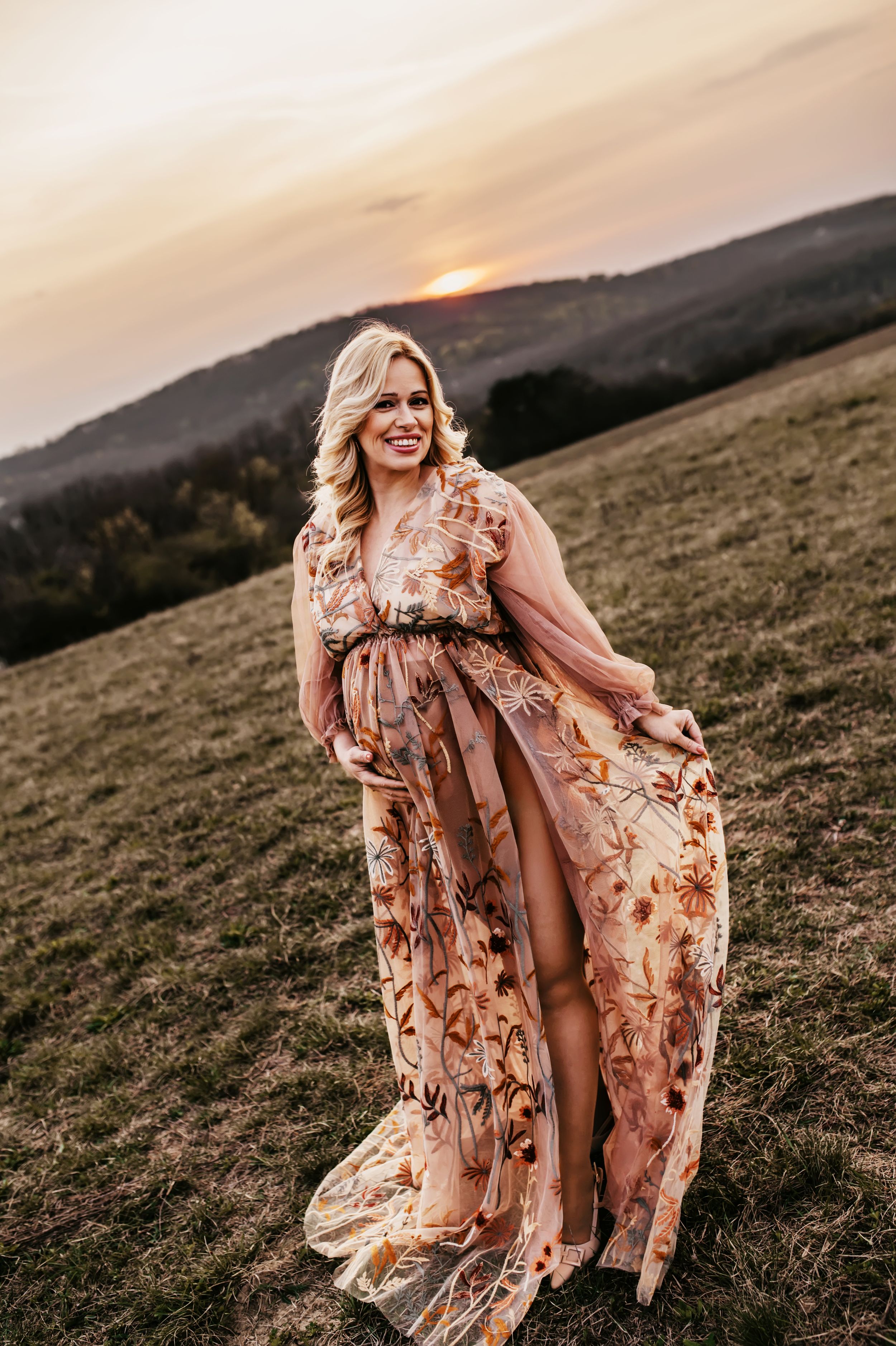 Pregnant woman in floral maxi dress poses on hillside at golden sunset, smiling gracefully.