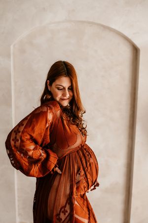 Expecting mother in a flowing rust-colored dress stands against a white wall in warm lighting.
