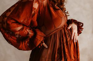 Close-up of a flowing rust-colored dress with gathered fabric details and bishop sleeves against neutral background.