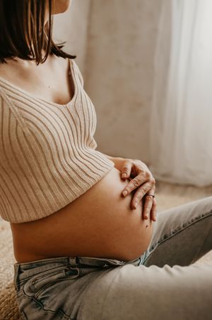 A pregnant woman in a cream ribbed sweater gently rests her hands on her exposed belly in soft window light.