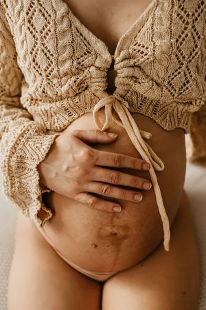 Delicate cream-colored knit sweater tied above pregnant belly with hands resting gently on bare stomach.