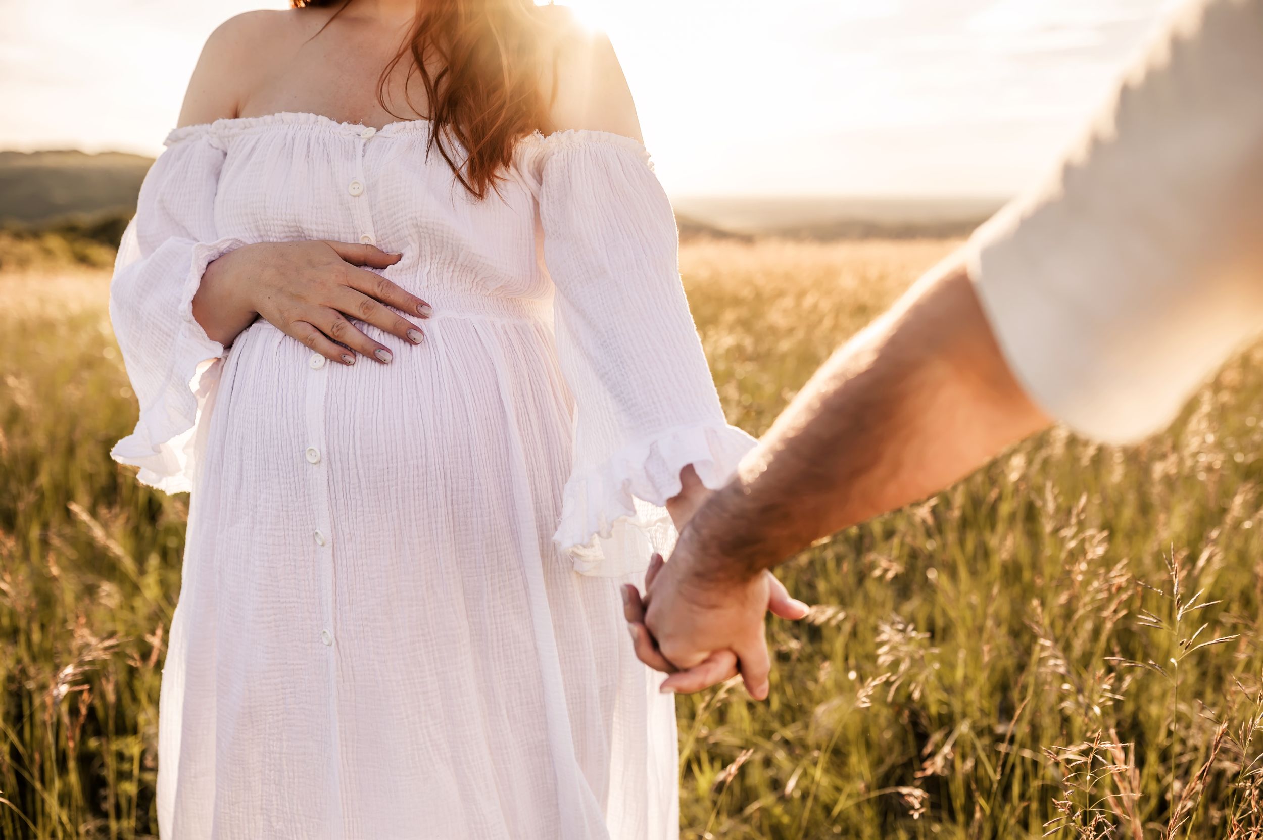 Pregnant woman in white dress holds hands with partner in golden wheat field at sunset.