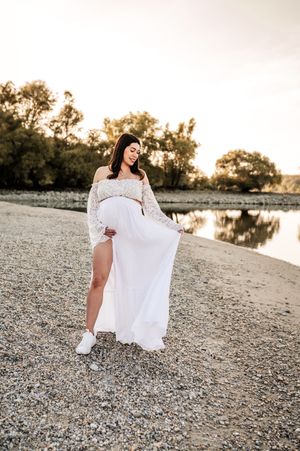 Ethereal maternity photo of an expectant mother in a flowing white lace dress standing by a serene lake at sunset.