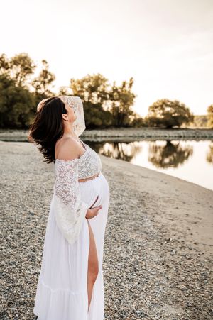 A pregnant person in a white lace maternity dress poses by a lake at sunset, creating a serene silhouette.