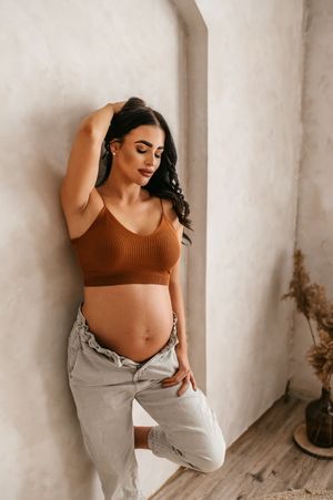 Beautiful maternity pose against white wall wearing brown crop top and light jeans in natural indoor lighting.