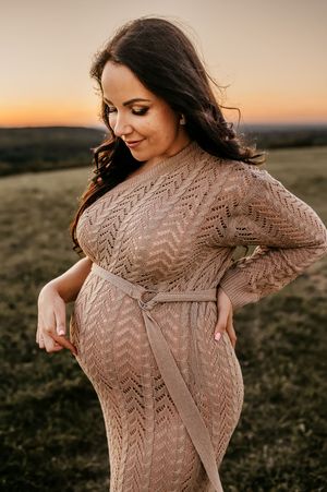 Expectant mother in knit sweater dress poses at sunset in golden field with warm autumn lighting.