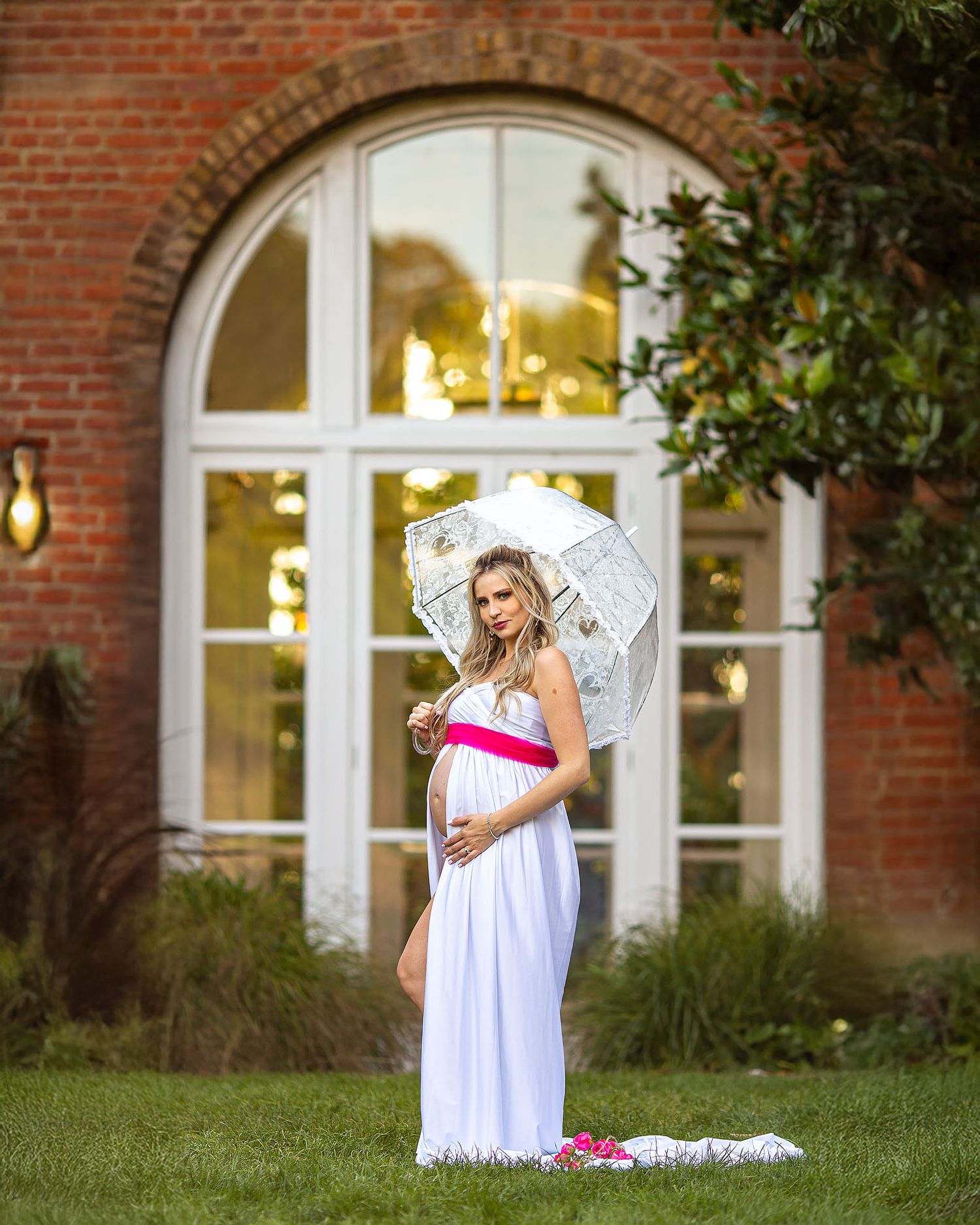 A pregnant person in a white dress stands in front of a brick building with arched windows at sunset holding a white parasol.
