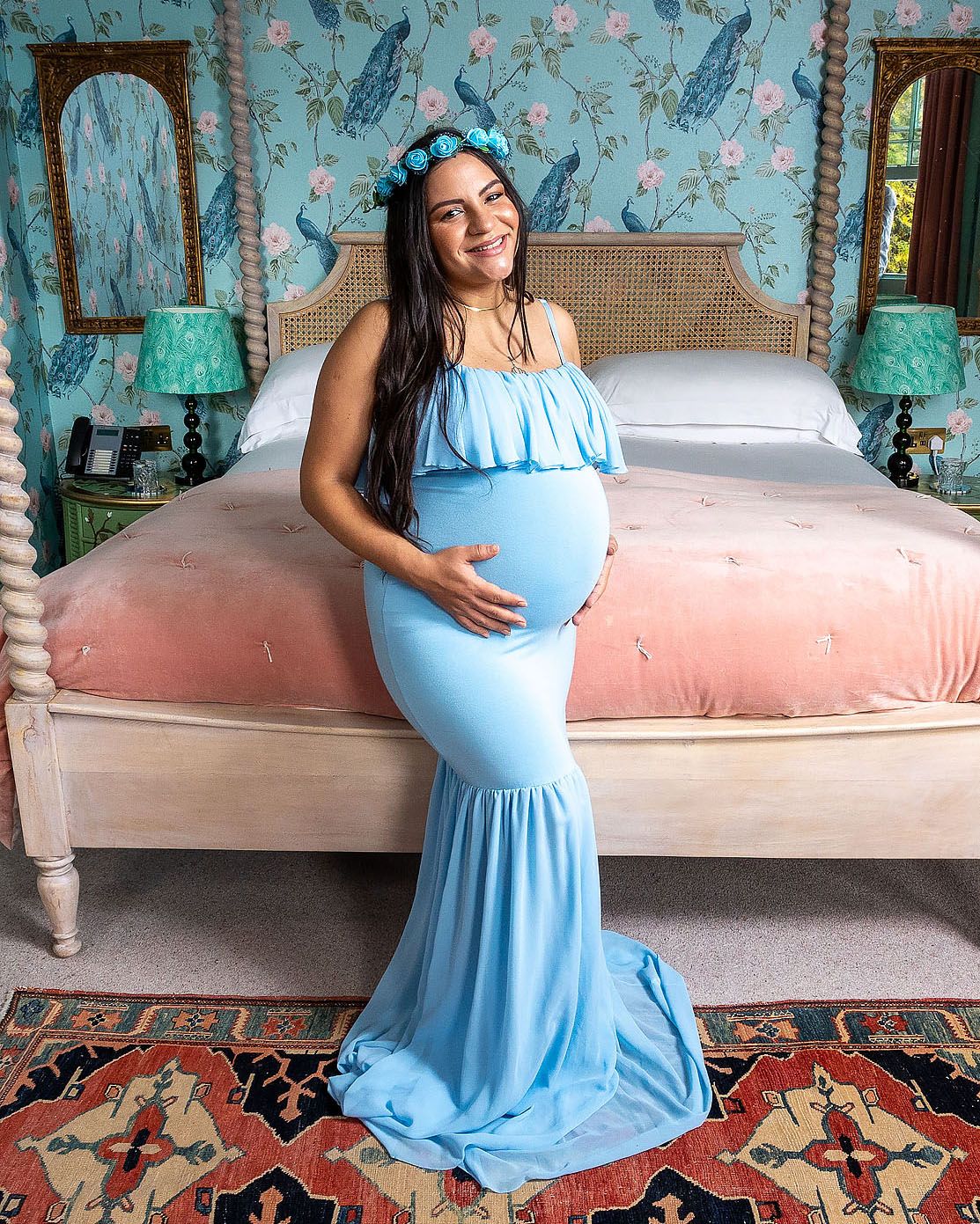 Expectant mother in flowing blue ruffle maternity gown poses in bedroom with floral wallpaper and vintage decor.
