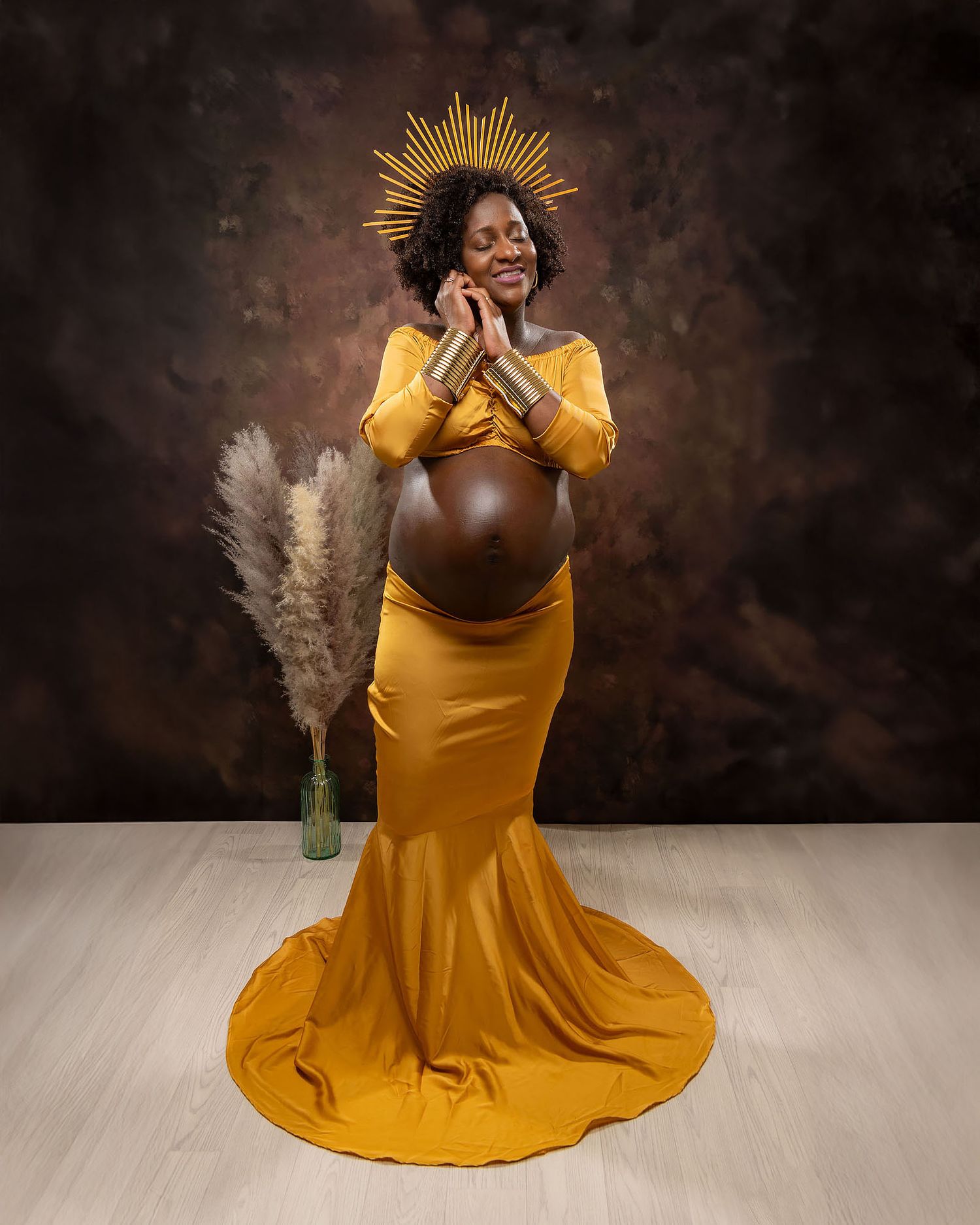 Maternity photo featuring a woman in a flowing mustard yellow gown with a golden sunburst crown against a dark backdrop.