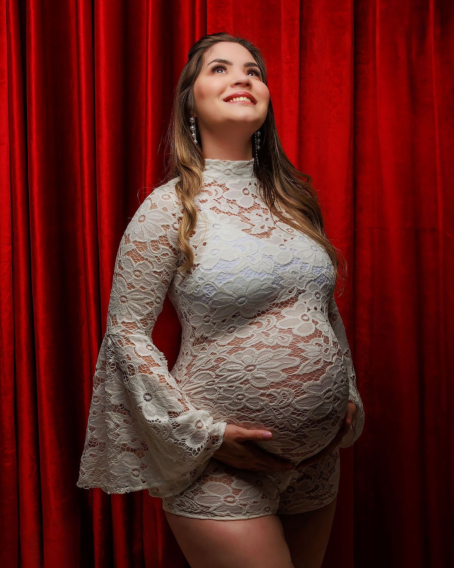 Expectant woman in white lace dress poses against red velvet curtain backdrop during maternity photoshoot.