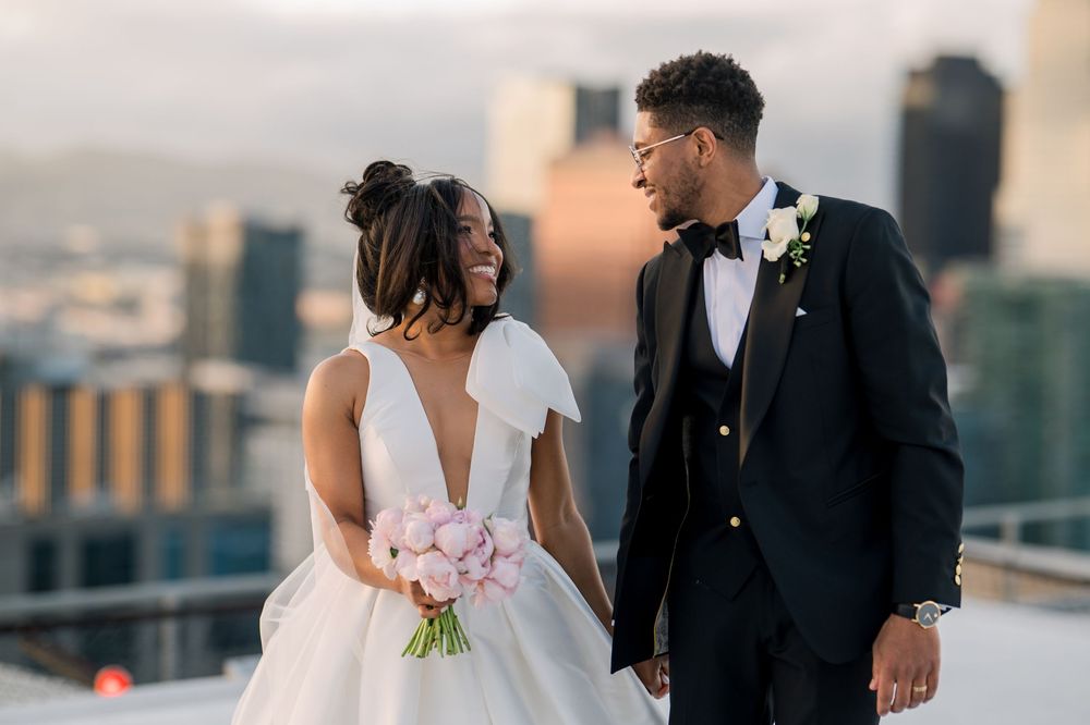 A newlywed couple sharing a romantic moment on a rooftop at sunset with a city skyline backdrop.