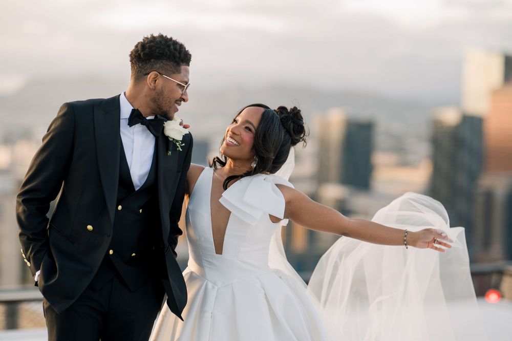 A joyful wedding couple laughs together on a city rooftop at sunset with urban skyline in the background.