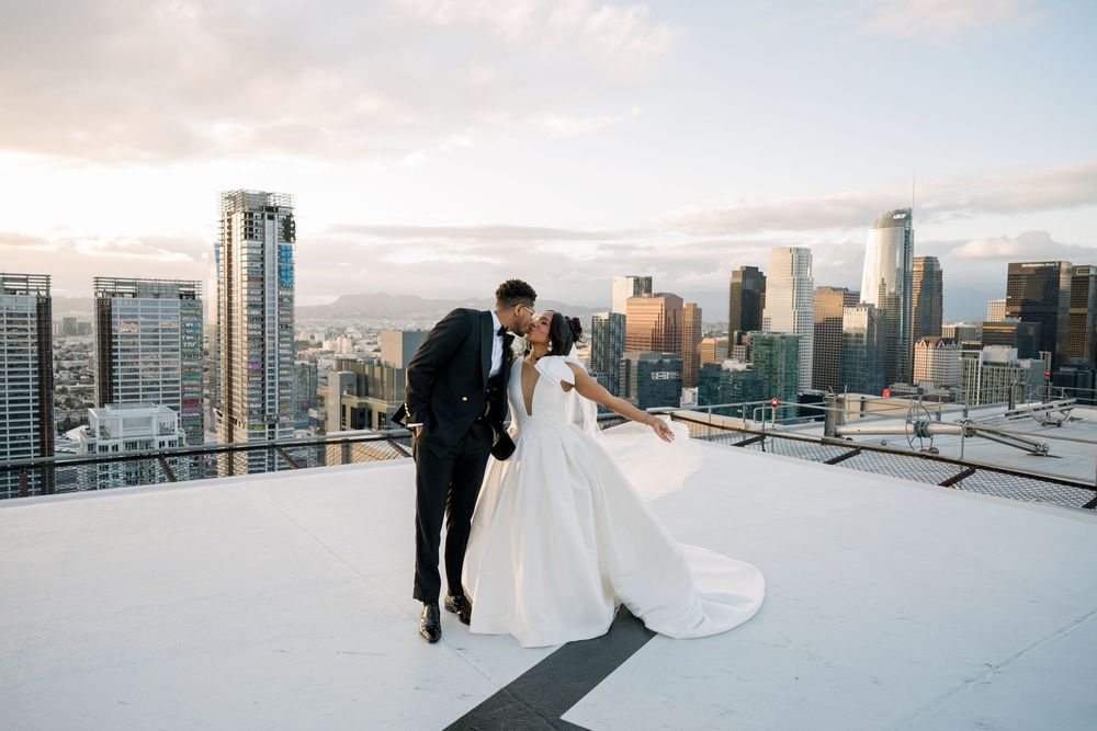 A couple in wedding attire share a romantic moment on a snow-covered rooftop with a dramatic city skyline backdrop.