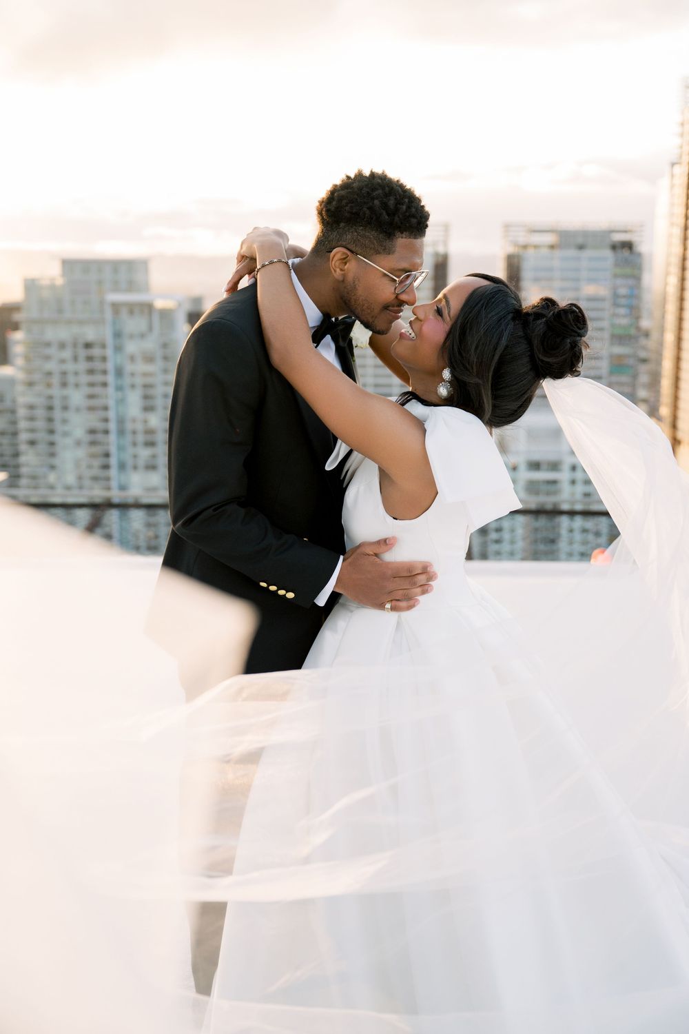 A romantic rooftop wedding moment with flowing white veil against a city skyline backdrop at sunset.