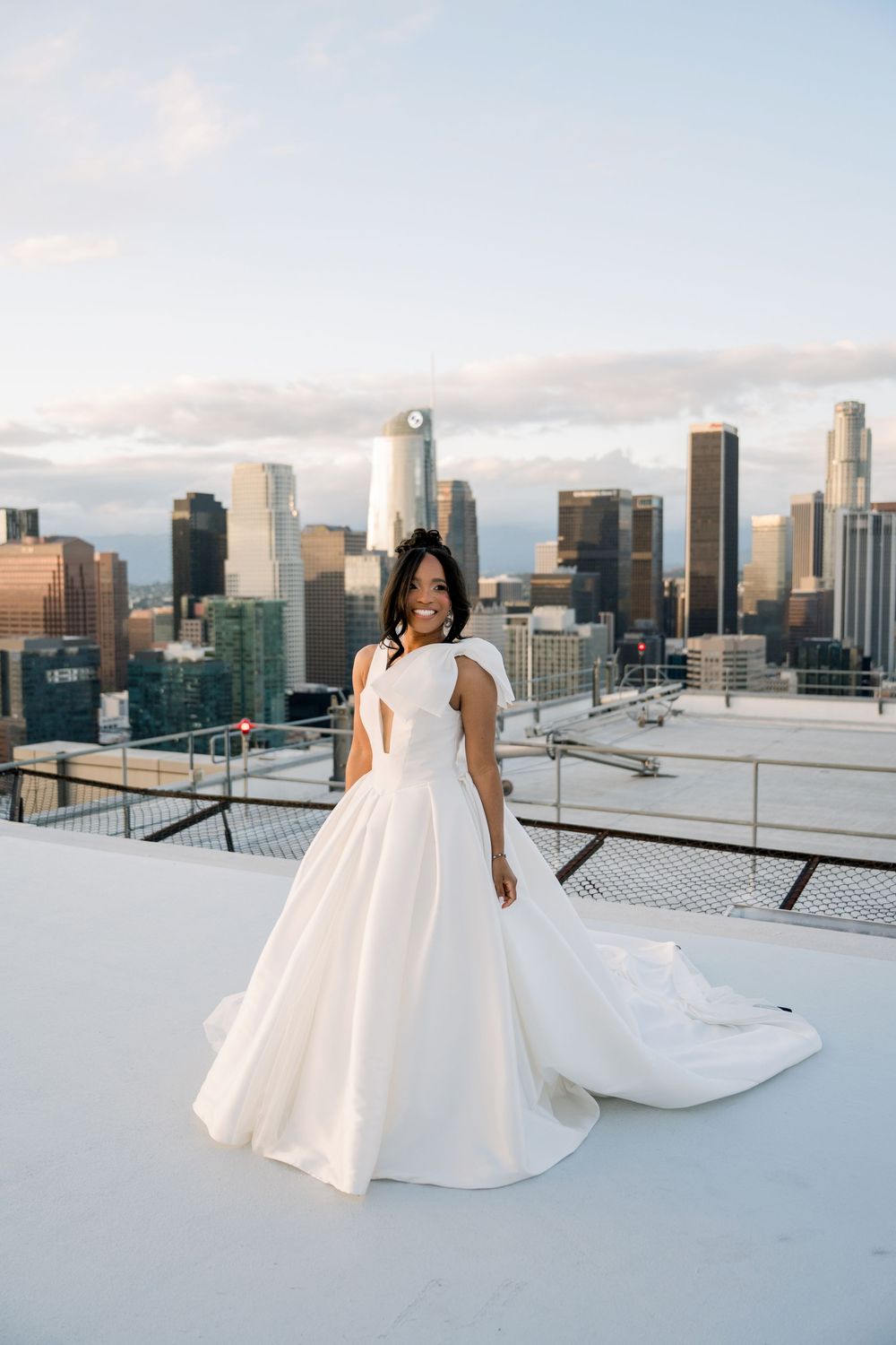 Elegant wedding dress with flowing train photographed on a rooftop against the Los Angeles city skyline at sunset.