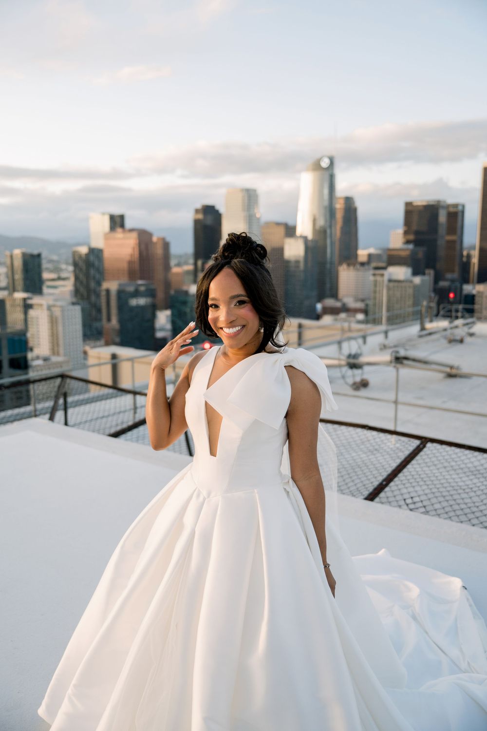 A person in an elegant white wedding gown poses on a rooftop with a cityscape backdrop at sunset.