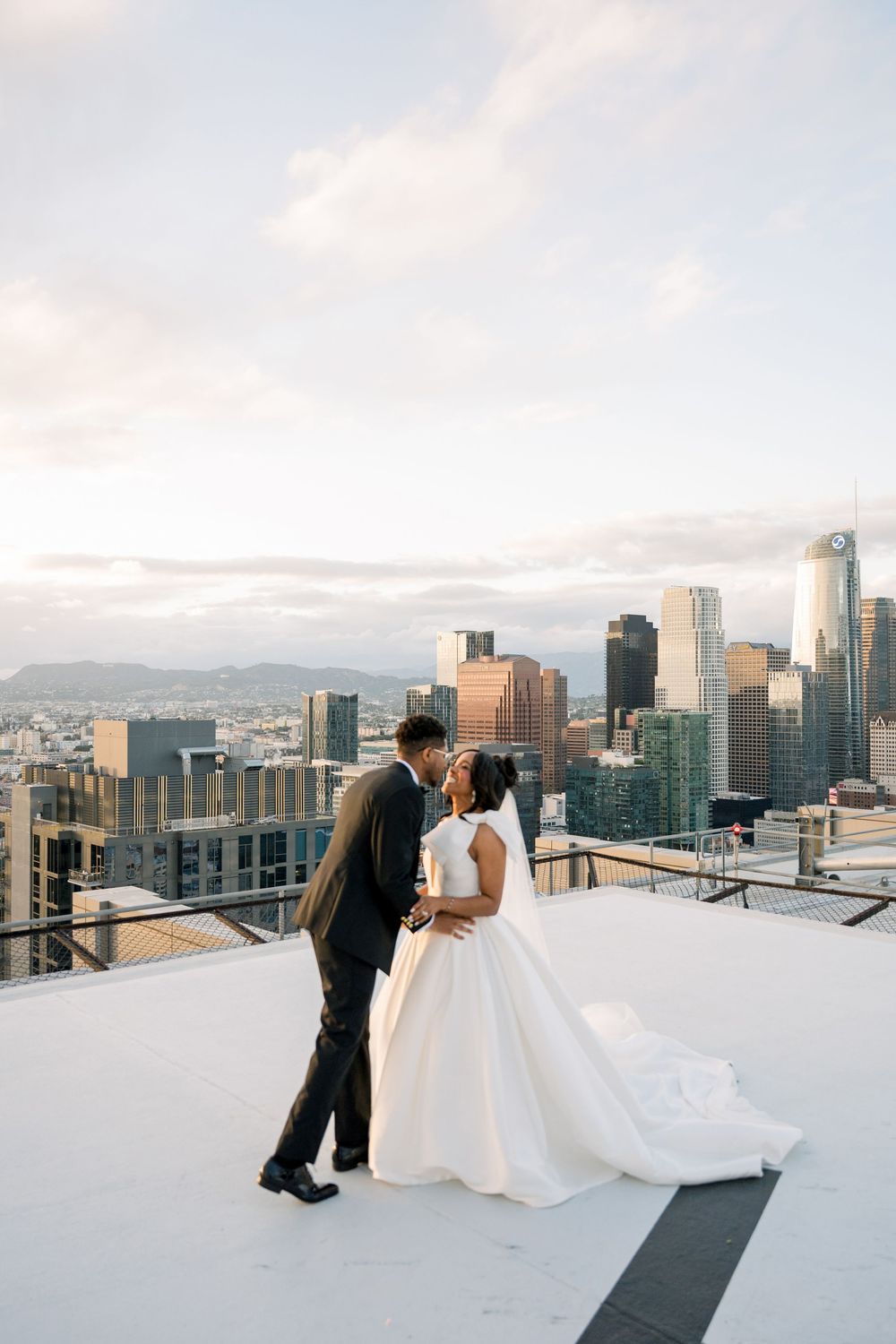 A couple shares an intimate moment on a rooftop overlooking the Los Angeles skyline at sunset.