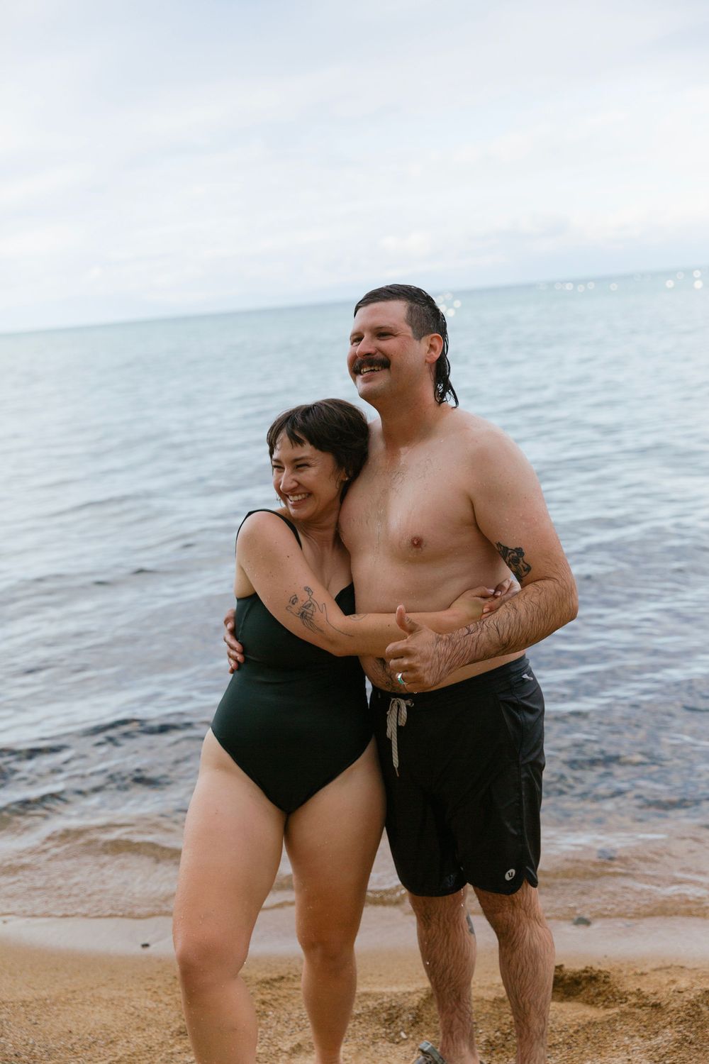 A joyful couple embraces while standing in swimwear on a beach with the ocean in the background.