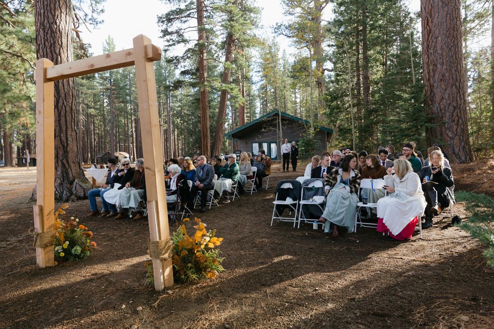 Rustic outdoor wedding setup in a pine forest with rows of white chairs and wooden arch.