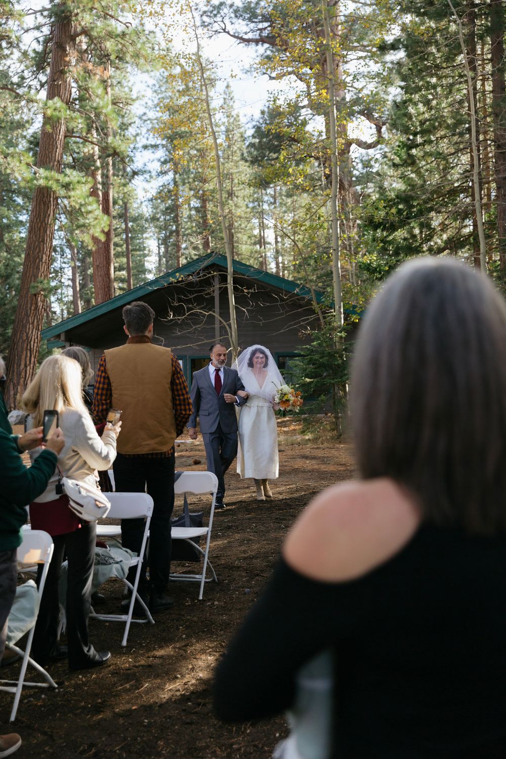 Wedding ceremony taking place in a secluded forest setting with guests seated on white chairs.