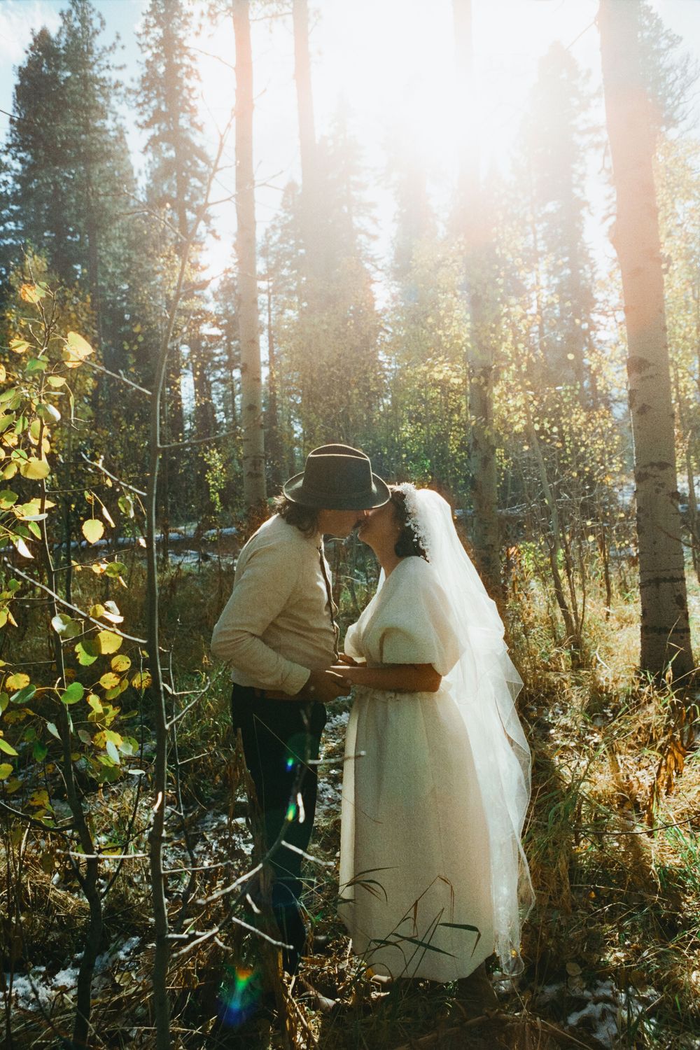A romantic forest wedding photo with sunlight streaming through pine trees as a couple embraces.