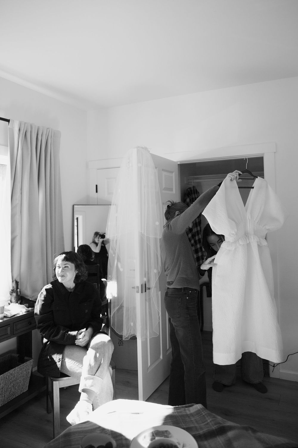 A black and white photo showing preparations in a room with a white dress hanging.