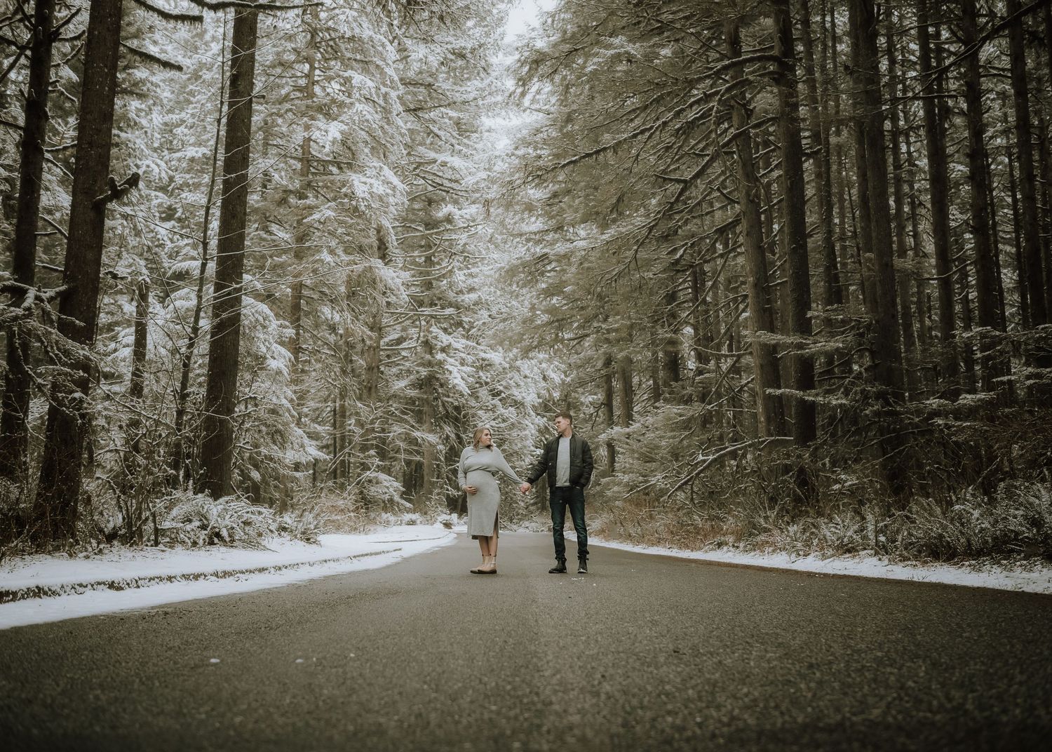 Two figures walk hand in hand down a snowy forest road lined with tall evergreen trees in winter.