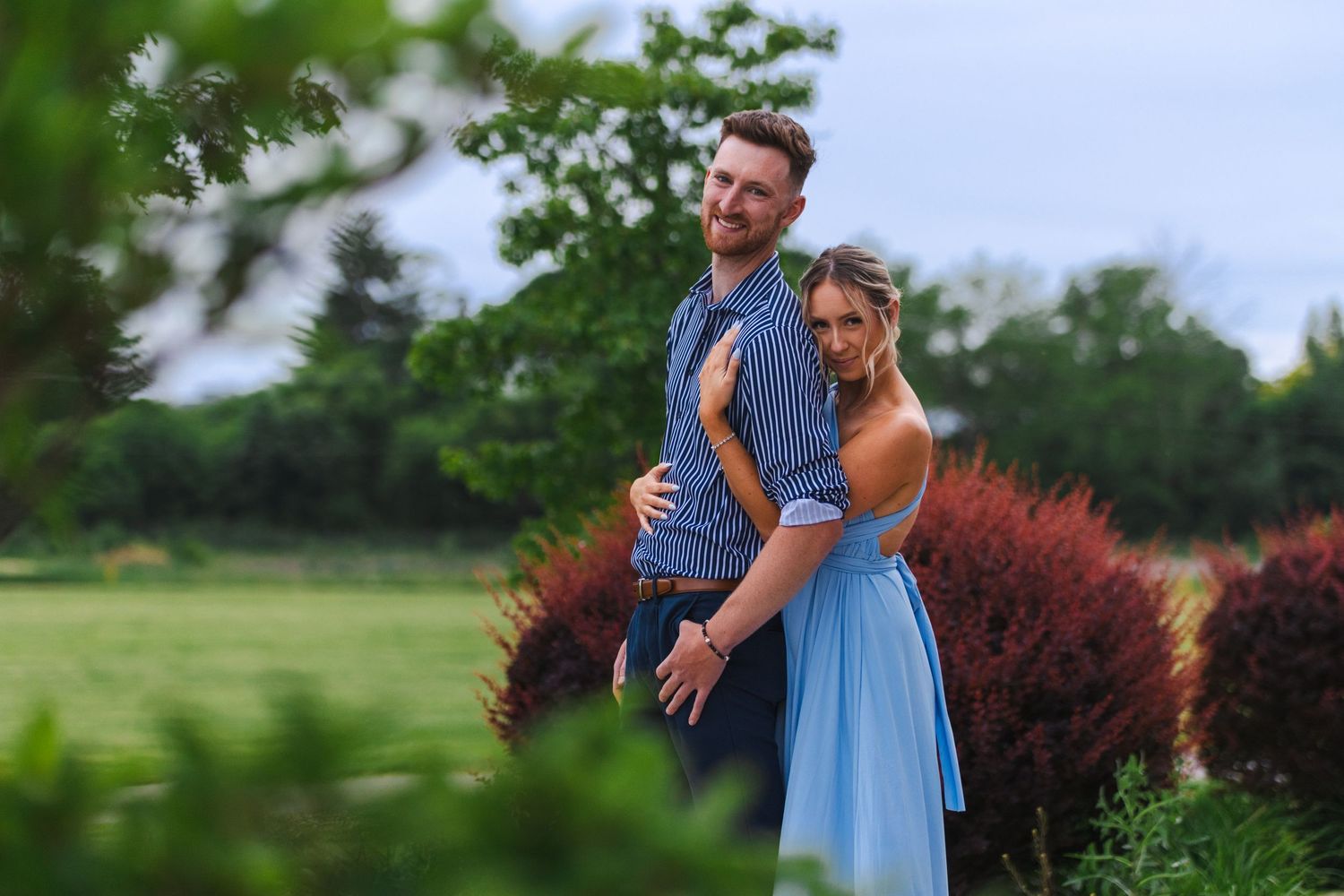 Couple in casual attire posing against blurred garden background.
