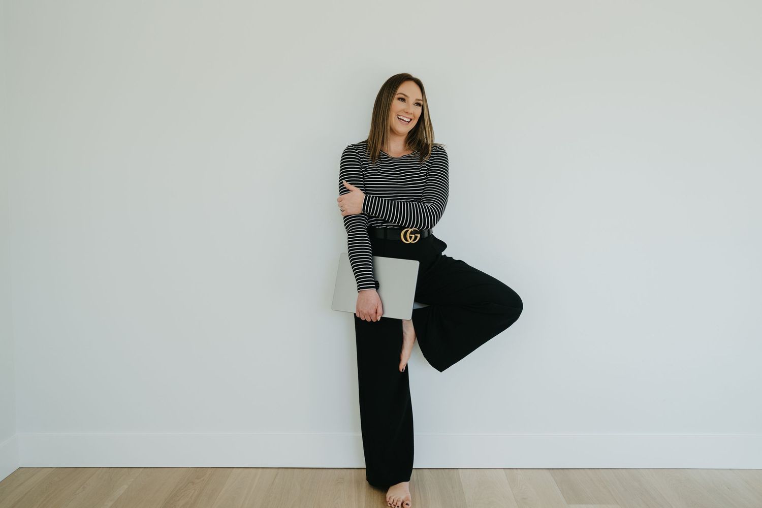 Person in casual gray sweater and black wide-leg pants poses with lifted leg against white wall.