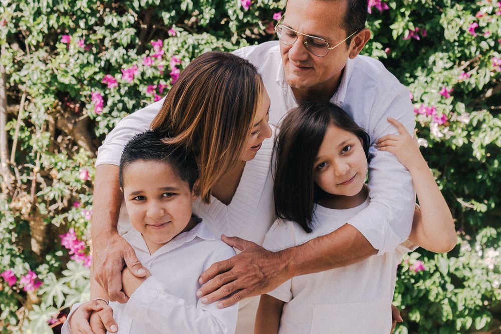 Family wearing white shirts embraces in front of blooming pink flowers in a garden setting.