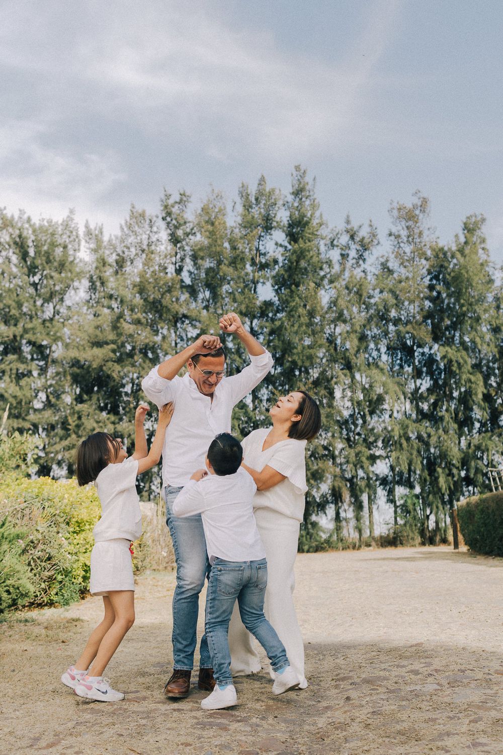 Family of four wearing white outfits playing and laughing together in front of tall trees on a sunny day.