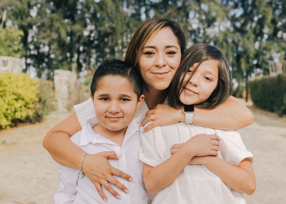 Three family members wearing white outfits embrace while posing outdoors with trees and greenery in the background.