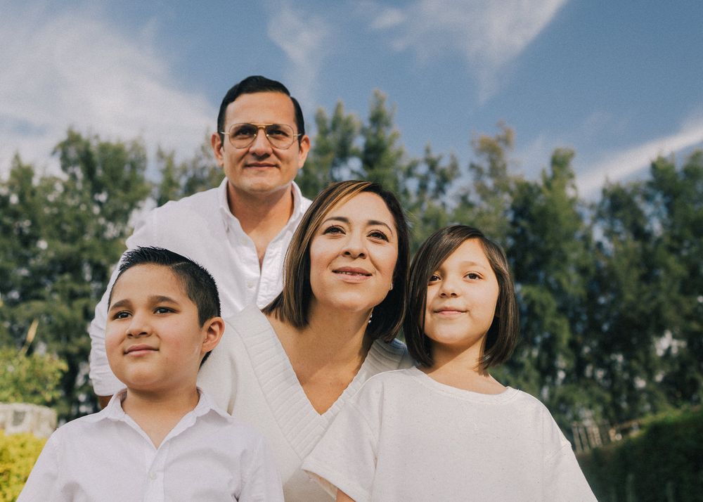 A close family group wearing white clothes poses together outdoors against a backdrop of green trees on a sunny day.