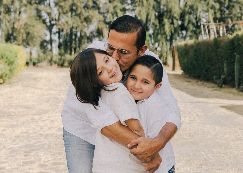 Three people wearing white shirts embrace warmly on a tree-lined dirt path on a sunny day.